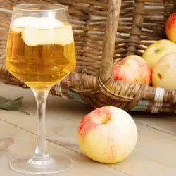 Image of a wine glass of hard cider with slices of apples floating in the glass, background of apples in a wicker basket.